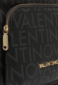 Bolsa de cuero negro texturizado con un patrón de logo repetido, cremallera en tono dorado y herrajes con un acabado brillante.