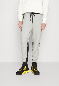 Grijze sweatpants met zwarte zijkanten, een touwlistriem in de taille en ribbels bij de manchetten, gecombineerd met zwart en gele sneakers. De stof is een textuur van katoenen mix.