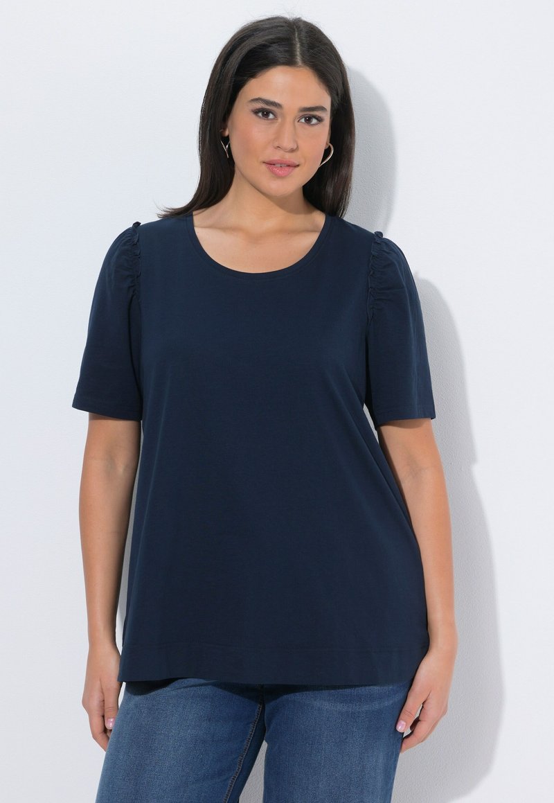 Mujer de cabello oscuro que lleva una camisa azul marino de manga corta con hombros fruncidos y vaqueros azules, de pie frente a un fondo blanco.