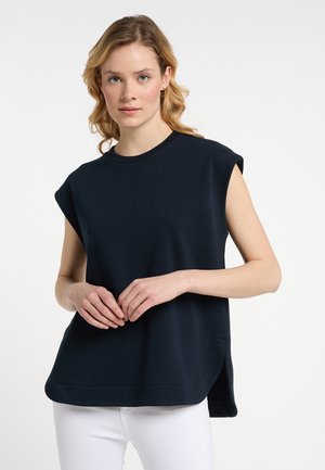 Vrouw met golvend blond haar die een mouwloos zwart topje en witte broek draagt, staand met handen zachtjes gevouwen.