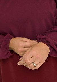 Burgundy long-sleeve dress with a textured fabric. Features gathered cuffs and smooth neck, hands clasped with a silver ring visible.