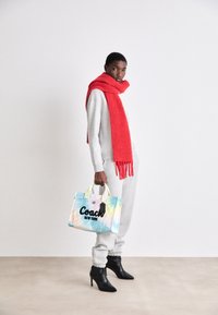 Sweat-shirt gris et jogging assorti avec une écharpe rouge vive. Bottes noires à talons hauts. Tenant un sac fourre-tout Coach multicolore avec logo noir.