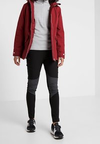 Červená izolovaná bunda s kapucí, šedý dlouhý rukáv a černé kalhoty s texturovanými detaily. Černé sportovní boty doplňují outfit.