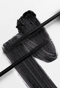 Scovolini per mascara neri con setole fini posati su una striscia nera spessa e lucida su sfondo bianco. Texture liscia e finitura ad alto lucido.