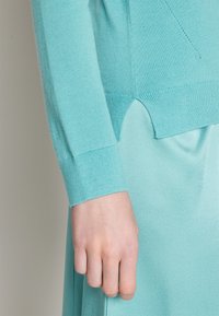 Gros plan d'une main posée à côté d'un pull en maille turquoise clair à manches longues avec poignets côtelés et jupe assortie.