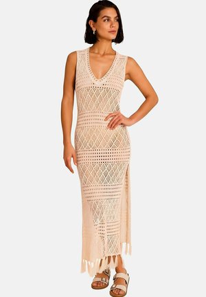 Femme portant une robe maxi beige en crochet sans manches avec des motifs géométriques et des fentes latérales, associée à des sandales blanches ouvertes.