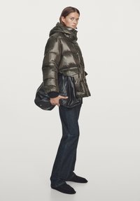 Kobieta w błyszczącej oliwkowej kurtce puchowej, ciemnych prostych dżinsach, czarnych butach, trzymająca dużą czarną skórzaną torbę, stojąca na jednolitym tle.