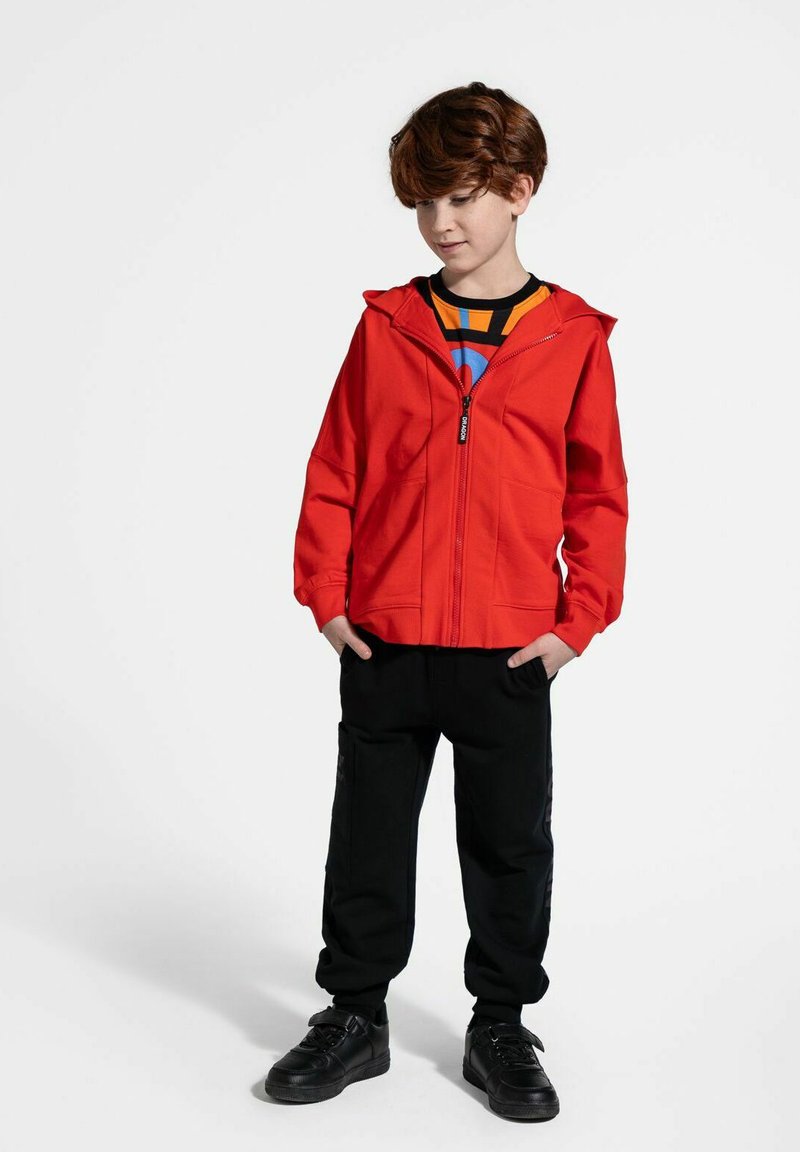 Czerwona kurtka z kapturem i zamkiem błyskawicznym, czarne spodnie i czarne sneakersy. Kurtka ma gładką teksturę i przednie kieszenie.