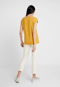 Senapsgul topp med korta ärmar, åtsittande design, mjuk textur, i kombination med vita nedklippta byxor och ljusgrå sneakers, sedd bakifrån.