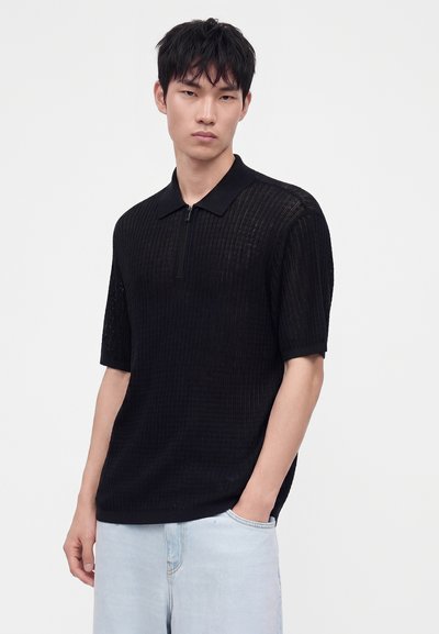 Jeune homme en polo noir texturé à manches courtes et jean bleu clair, debout avec une main dans la poche sur un fond uni.