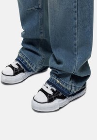 Sneaker basse in bianco e nero con superficie testurizzata, abbinate a jeans di denim blu oversize con orli sfilacciati e vestibilità ampia.