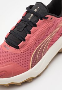 Zapatilla deportiva rosa con parte superior de malla, cordones negros y acentos dorados. Presenta un diseño texturizado y una suela de goma con agarre.