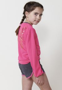 Chemise rose à manches longues avec un graphique d'ailes métalliques dans le dos, associée à un short gris avec des bordures roses. Tissu lisse, coupe décontractée.