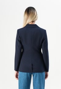 Žena s dlouhými blond vlasy, oblečená v přiléhavém tmavě modrém saku a modrých džínách, stojící zády k našedlému pozadí.