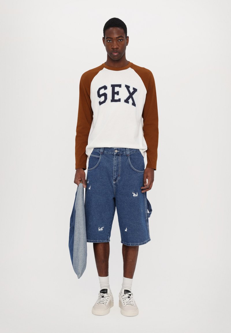 White long-sleeve shirt with brown raglan sleeves and navy lettering, paired with blue denim shorts featuring embroidered rabbit designs.