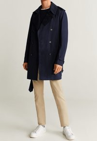 Mężczyzna w granatowym płaszczu dwurzędowym, beżowych spodniach, czarnej koszuli i białych trampkach na jednolitym jasnym tle.