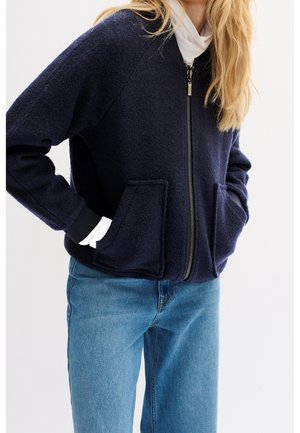 Personne portant une veste zippée bleu marine avec de grandes poches à l'avant, une chemise blanche en dessous et un jean bleu sur un fond uni.