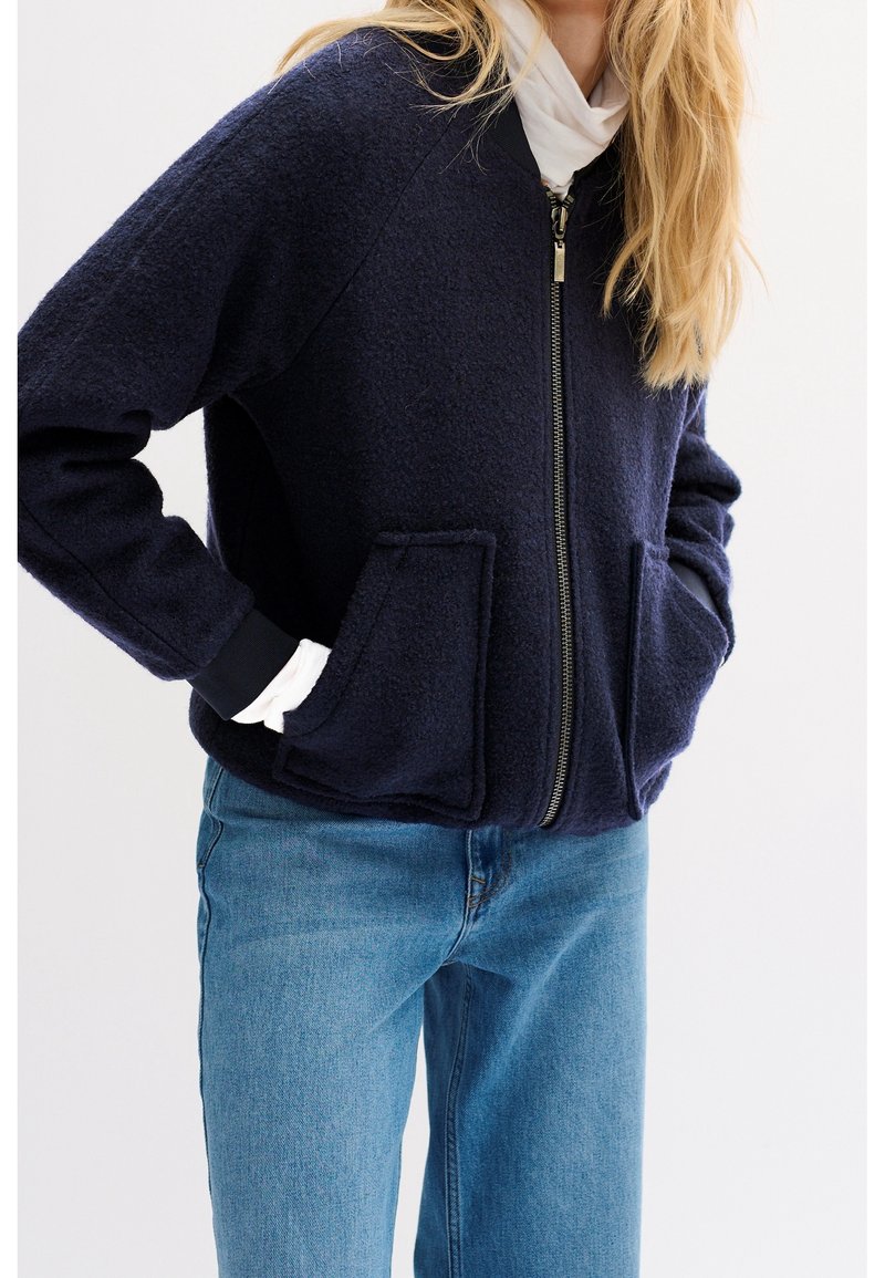 Personne portant une veste zippée bleu marine avec de grandes poches à l'avant, une chemise blanche en dessous et un jean bleu sur un fond uni.
