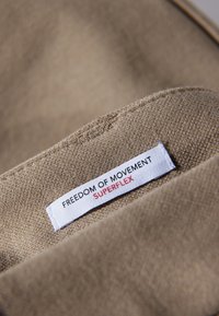 Beżowa tkanina z białą tkankową metką z czarnym napisem "FREEDOM OF MOVEMENT" i czerwonym napisem "SUPERFLEX".