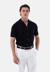 Polo shirt corto blu scuro a maniche corte con colletto button-down, abbinato a pantaloni bianchi con pinces e una cintura in pelle marrone.