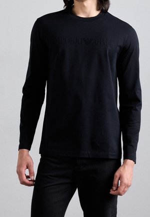 Mann trägt ein schwarzes langärmeliges Emporio Armani Shirt mit dezentem Logo-Schriftzug auf der Brust, kombiniert mit schwarzen Hosen.