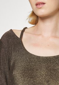 Glänzendes Bronzeoberteil mit schulterfreiem Design, strukturiertem Stoff und rundem Ausschnitt. Trägerdetails sind auf der linken Schulter sichtbar.