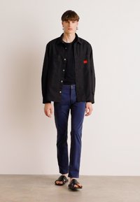 Cămașă neagră cu butoni, tricou negru, pantaloni bleumarin și sandale negre. Texturi netede, croială dreaptă și accent roșu pe cămașă.