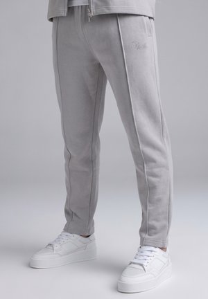Pantalones deportivos grises con costuras laterales, llevados con zapatillas blancas de corte bajo sobre fondo neutro.