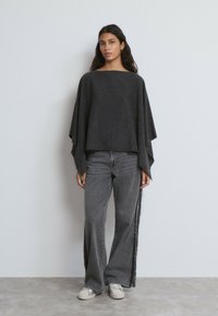 Top tipo poncho de gran tamaño en gris oscuro, con un escote amplio y mangas sueltas, combinado con jeans acampanados grises que tienen aberturas laterales.