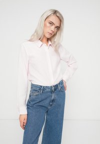 Camicia button-up rosa chiaro con maniche lunghe e una texture a righe, abbinata a jeans in denim blu a vita alta. Design minimalista, senza motivi visibili.