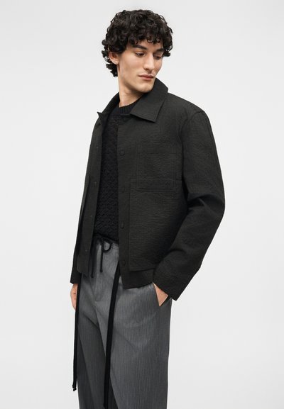 Άνδρας με κατσαρά μαλλιά που φοράει υφασμάτινο μαύρο σακάκι, μαύρο πουλόβερ με σχέδια και γκρι παντελόνι με λεπτές ρίγες και μαύρα κορδόνια.