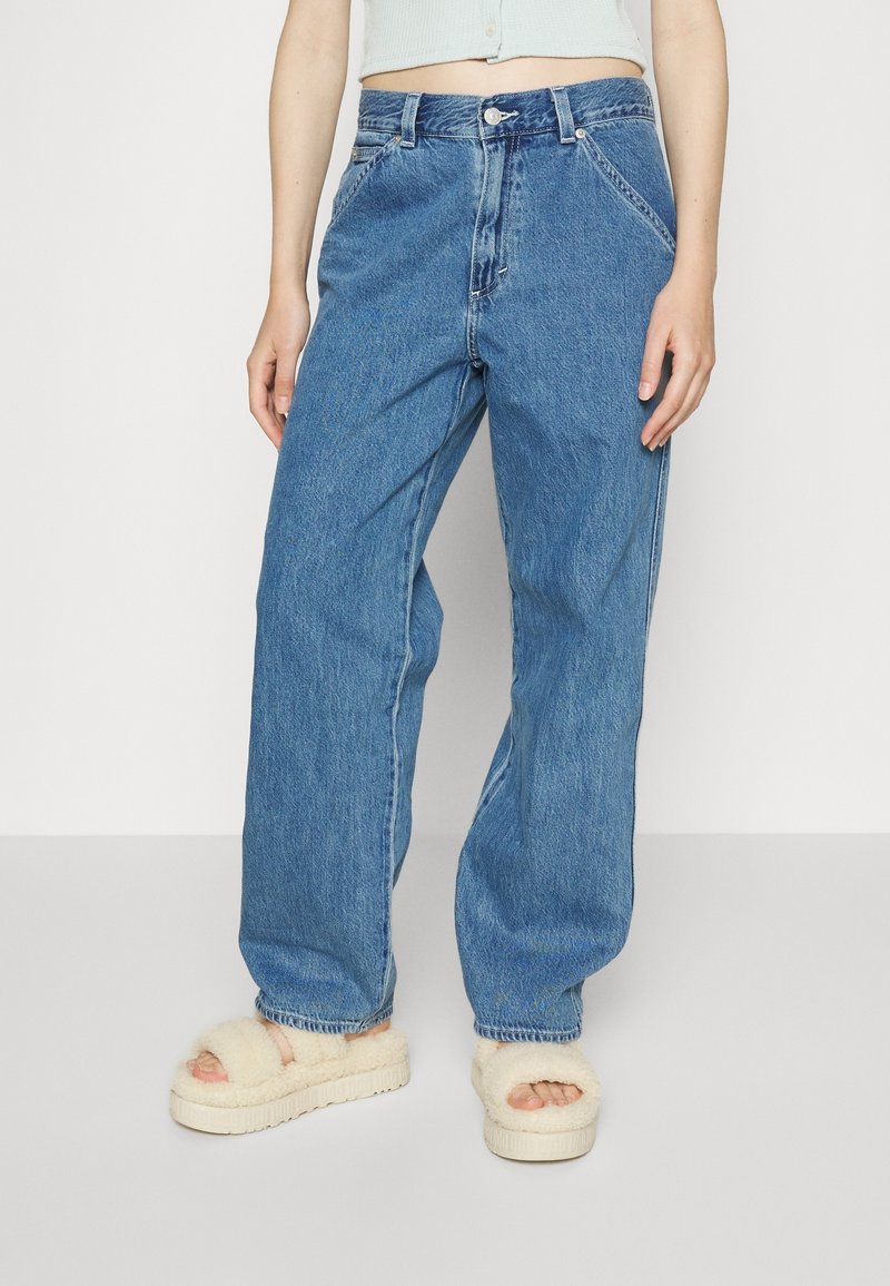 Torso inférieur portant un jean taille haute décontracté bleu clair et des sandales plateformes crème en peluche sur fond blanc.