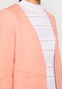 Blazer rose pâle à la texture lisse et au design ajusté, superposé à un haut blanc à col montant avec des détails tricotés horizontaux à motif.
