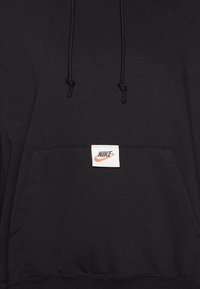 Melna kapuce ar priekšējo kangaroo kabatu, regulējamām auklām un baltu etiķeti, kurā ir sarkans un melns Nike logotips. Mīksta auduma tekstūra.