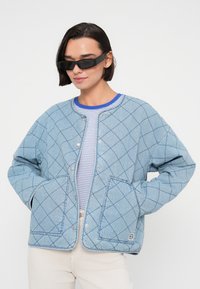 Γαλάζιο καπιτονέ τζιν μπουφάν με αντίθετη μπλε ραφή, κουμπιά με τσάκισμα και δύο τσέπες μπροστά· φορεμένο πάνω από ριγέ τοπ.