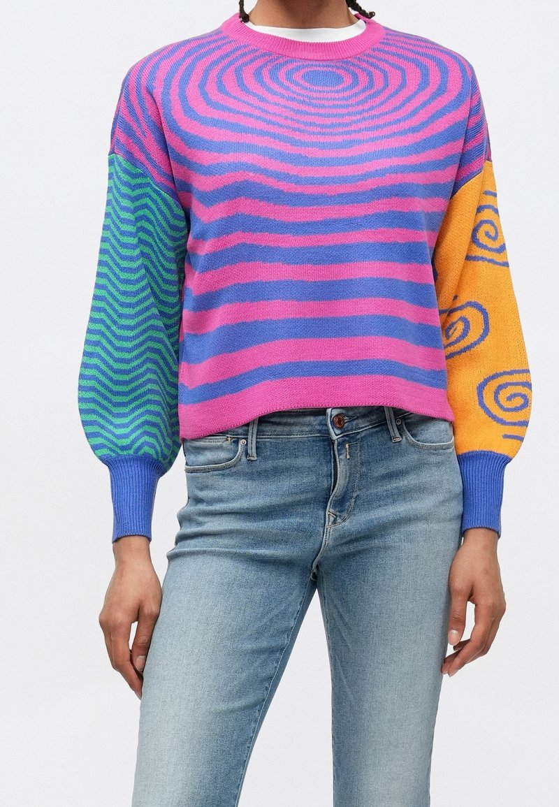 Femme portant un pull coloré avec des rayures concentriques roses et violettes sur le corps, une manche gauche verte en zigzag et une manche droite orange avec des tourbillons bleus.