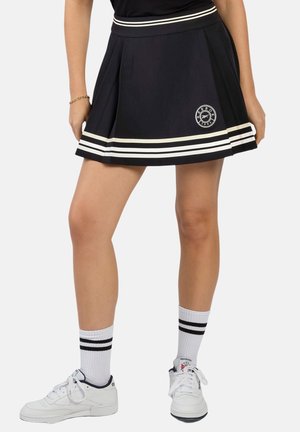 Mujer con falda negra plisada con rayas blancas y logo, calcetines blancos con rayas negras y zapatillas blancas, de pie con los pies separados.