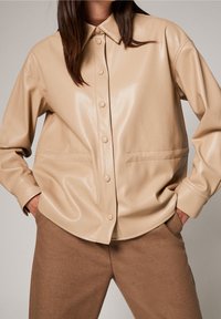 Veste chemise en simulateur cuir beige avec un front à boutons, un col pointu et des poches latérales ; texture lisse et coupe décontractée. Pantalons marron présentés.
