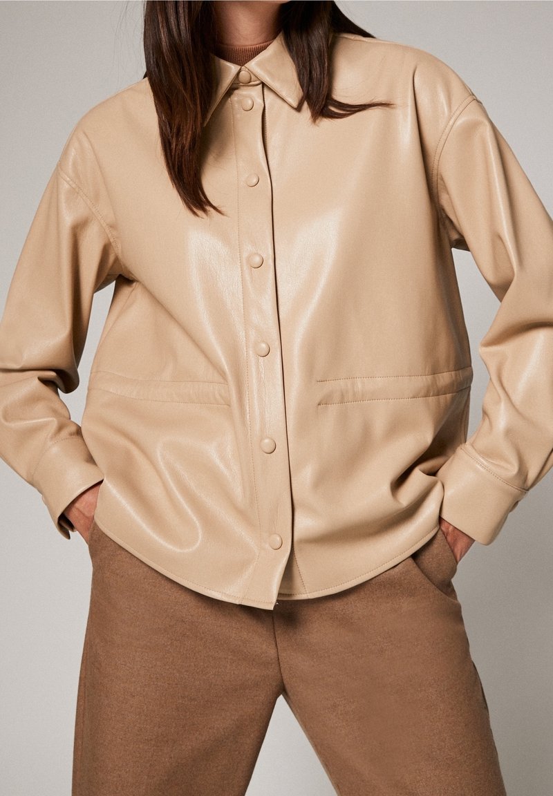 Veste chemise en simulateur cuir beige avec un front à boutons, un col pointu et des poches latérales ; texture lisse et coupe décontractée. Pantalons marron présentés.