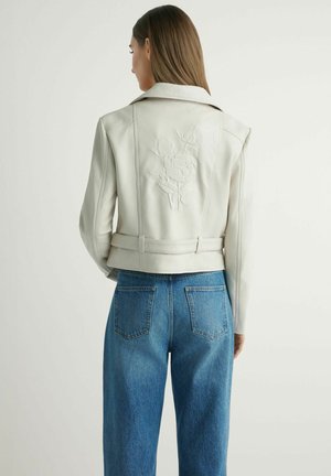 Kobieta w białej skórzanej kurtce z tłoczonym wzorem kwiatowym na plecach, ubrana w niebieskie dżinsy z wysokim stanem, stojąca tyłem.