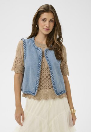 Nainen, joka pukeutunut beigeen pitsiseen lyhythihaiseen toppiin, vaaleansiniseen farkkulestiin, jossa punotut reunat, kerrokselliseen kermaan vivahtavaan hameeseen, kultainen kalvosinrannekoru ja renkorenkaankorvakorut.