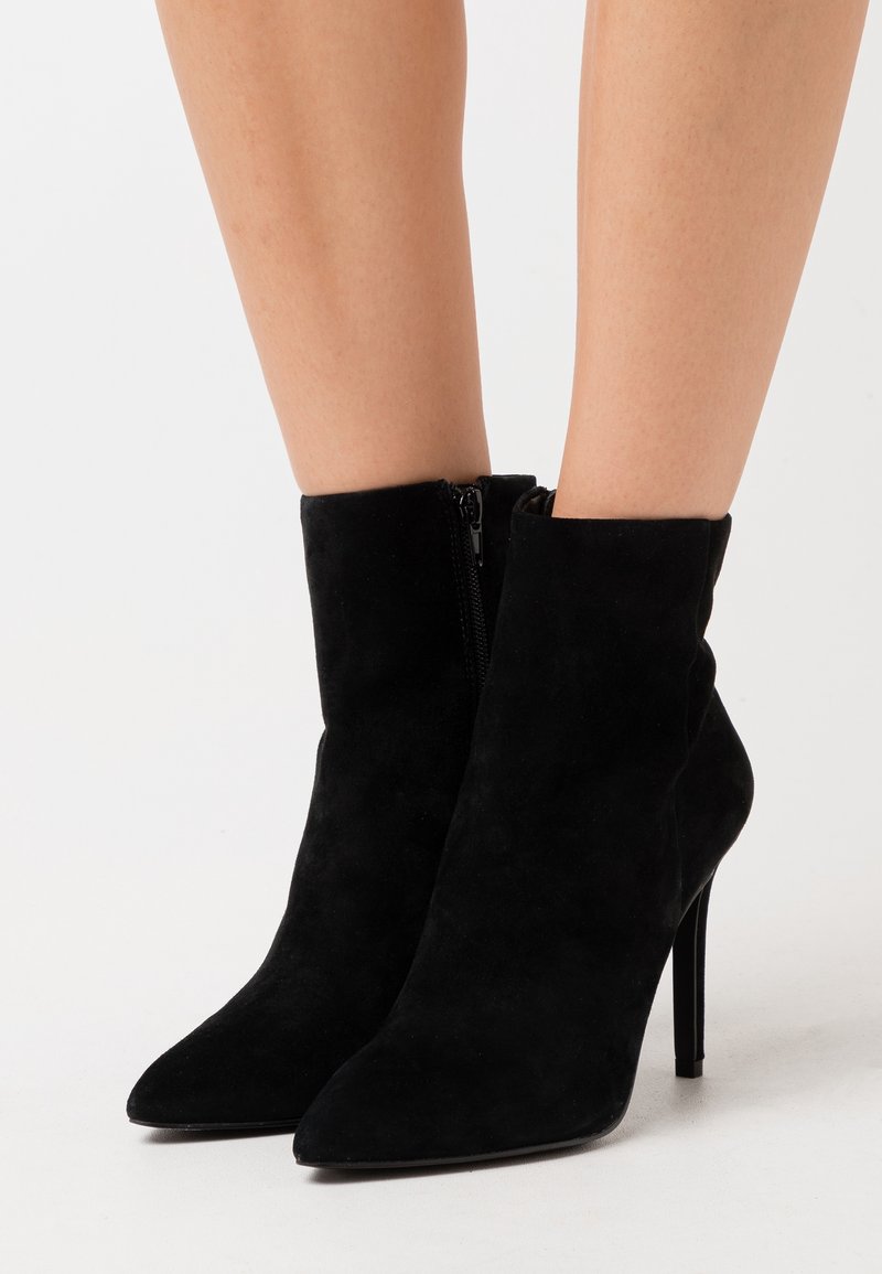 Bas noir en daim portant des bottines à talons hauts avec bouts pointus et fermetures éclair latérales, portées sur des jambes nues.