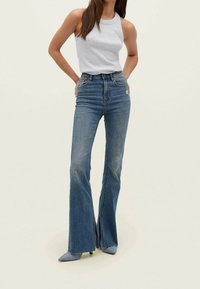 Jeans acampanados de color azul claro con cintura alta, acabado desgastado y dobladillo deshilachado. Combinados con una blusa sin mangas gris y zapatos de punta azul claro.