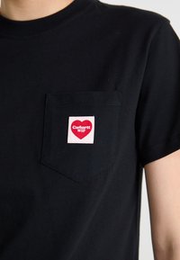 Schwarzes T-Shirt aus Baumwolle mit einer Brusttasche auf der linken Seite, die ein quadratisches Etikett in Pink und Rot trägt, auf dem "Carhartt WIP" steht.