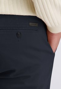 Marineblauwe broek heeft een gestructureerde afwerking, een enkele knoopsluiting en een klein merkembleem op de achterzak.