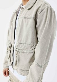 Grå denimjakke med lynlås, krave, to lynlåslommer samt elastiske manchetter og talje. Blød tekstur og afslappet pasform.