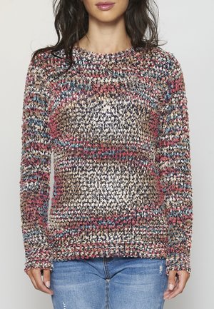 Kvinde iført en flerfarvet, struktureret strikket sweater med lange ærmer og blå slidte jeans, stående mod en ensfarvet baggrund.