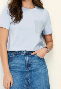 Vaaleansininen puuvillainen t-paita, jossa on lyhyet hihat ja teksturoitu tasku, jossa on sykerökuvio. Yhdistetty farkkuhameen kanssa.