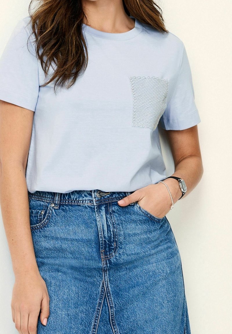 Vaaleansininen puuvillainen t-paita, jossa on lyhyet hihat ja teksturoitu tasku, jossa on sykerökuvio. Yhdistetty farkkuhameen kanssa.