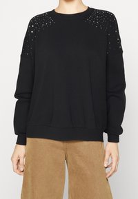 Sudadera negra con lentejuelas en los hombros, puños y bajo acanalados, y un corte relajado. Tejido suave con una textura lisa.